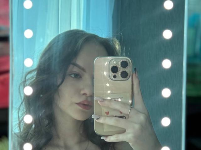 WeksaMur, com cabelos castanhos ondulados, segurando um iPhone bege, tirando uma selfie no espelho com um fundo iluminado, mostrando seus lábios carnudos e unhas com pequenos corações vermelhos..