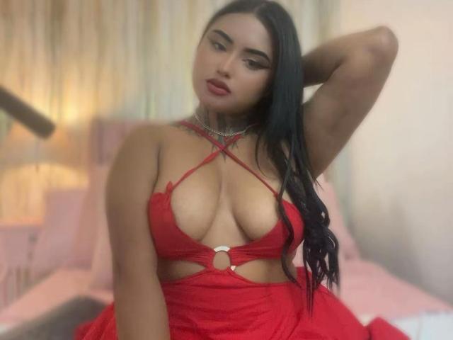 LairaBeamont in red lingerie, showing her big boobs, with a cutout top, long black hair, arm raised, looking down, red lipstick, in a blurry bedroom.