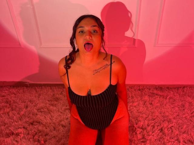IvyJoness kneeling on a pink shag carpet, tongue out, wearing a black pinstripe bodysuit with spaghetti straps, exposing her tits, with tattoos on her chest. Red lighting.