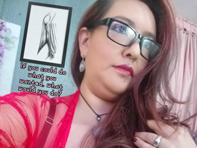TerraMegaBoobs com óculos, blusa vermelha de renda e cabelo castanho longo, usando brincos e um colar, segurando o cabelo, olhando para a câmera, com uma expressão confiante..