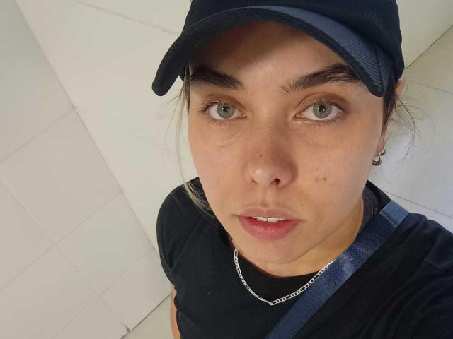 RosyG is in a close-up selfie with a neutral expression. She has light green eyes, dark eyebrows, and a small mole on her left cheek. She wears a dark blue cap, a black t-shirt, and a silver chain necklace. Her right shoulder has a blue strap. The image is clear and well-lit, focusing on her face. The background is white and slightly out of focus.