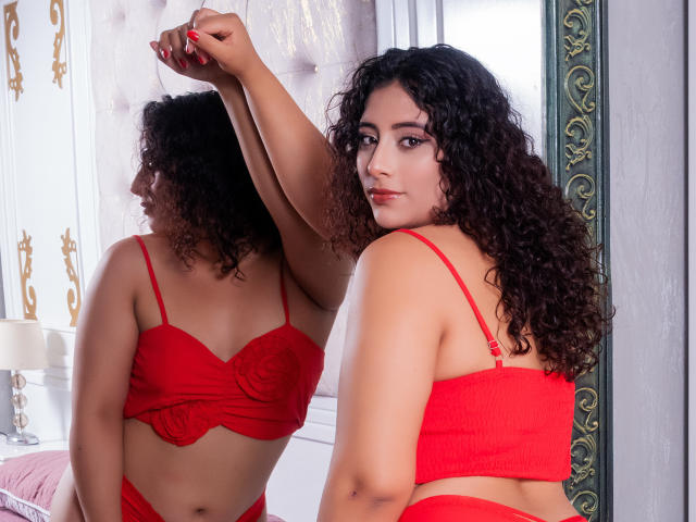 NicollWest with curly dark hair, red bra, and matching red underwear, reflects in a mirror, showing her back and looking over her shoulder with a slight smile.