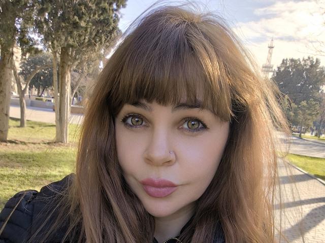 DiamondMature has brown, wavy hair with bangs, green eyes, a nose ring, and pink lipstick. She's wearing a black top, looking serious, with a grassy, tree-filled background.