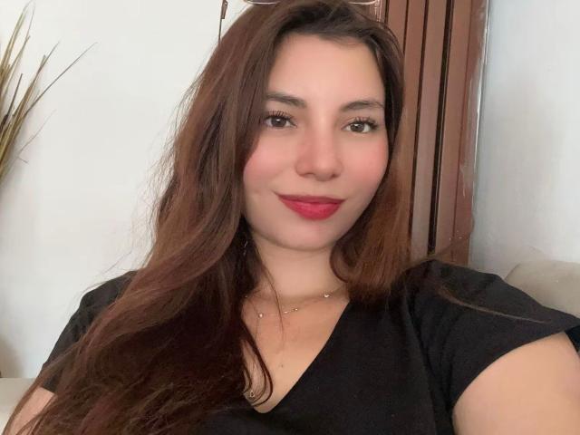LinaFoxyHot has long brown hair, pink lipstick, and is smiling with a black shirt on. She's wearing a small necklace and has a cute, attractive face.