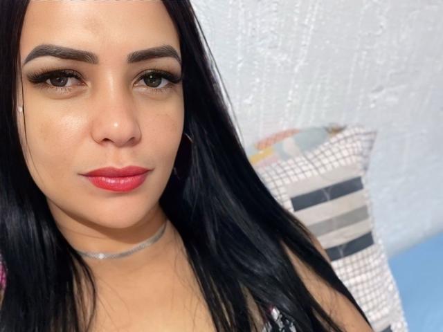 Lualy has long black hair, brown eyes with thick eyelashes, red lipstick, and a silver choker. She's looking slightly to the side with a subtle smile.