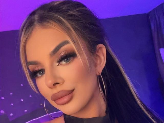 IvyRaven with blonde highlights, thick eyebrows, long eyelashes, dark lipstick, and large hoop earrings, looking at camera with a slight smile, blue-purple light in background.