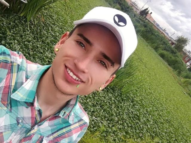 IamSmiith, ein junger Mann mit gebräunter Haut, kurzen braunen Haaren, einer weißen Mütze, grünen Ohrringen, einem Nasenring und einem karierten Hemd, lächelnd, Hintergrund: grünes Feld, bewölkter Himmel..