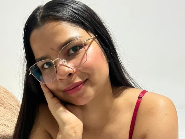 AmaiaSaenz with long black hair, red glasses, and red strap dress, touching her cheek, smiling, showing small acne on her nose, with a cute, soft expression.