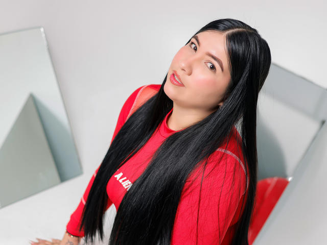 Alanah Wells mit langem, schwarzem Haar, einem roten Hemd und roten Lippen, die leicht zur Seite schaut und dabei ihre Brüste zur Schau stellt, sitzt in einem hellen Raum mit einem Spiegel..
