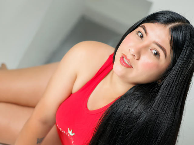 Alanah Wells, con lunghi capelli neri, canottiera rossa e lucido rosso sulle labbra, guarda la fotocamera, mostrando il seno, seduta con il sedere leggermente sollevato..