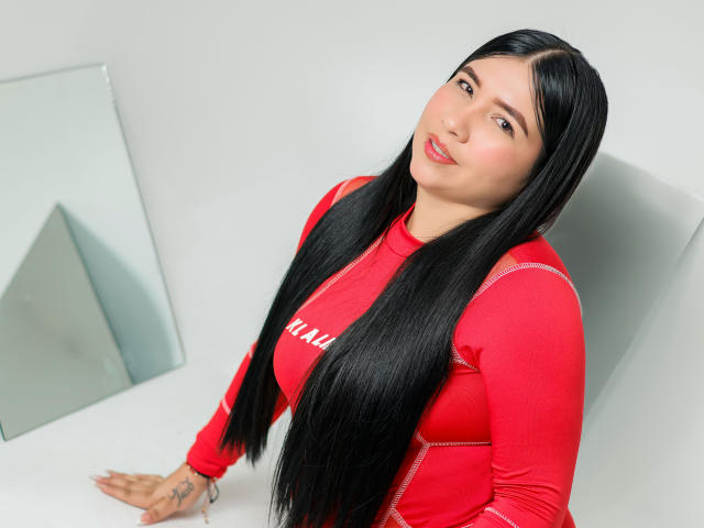 Alanah Wells, com cabelos longos e pretos, vestindo uma blusa vermelha de manga comprida, sentada com as mãos em uma superfície branca, sorrindo levemente, com batom rosa, tatuagem na mão direita, olhando para a câmera..