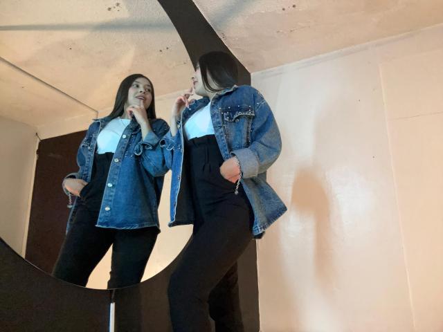 EmmaLombardi standing in front of a mirror, wearing a blue denim jacket, white tee, and black high-waisted pants, looking down with one hand near her mouth.