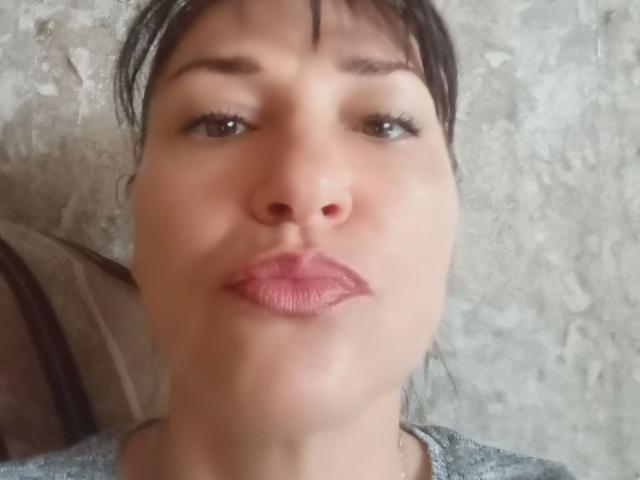 A photo of MonicaErotisch-hot with puffy pink lips, brown eyes, and dark hair, showing a slightly annoyed or bored expression, wearing a gray top.