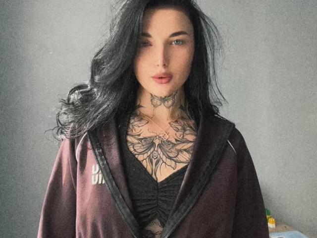 AlinkaMandarinka with black wavy hair, tattooed chest and neck, wearing a dark zip-up hoodie, looking at the camera with blue eyes and red lipstick.