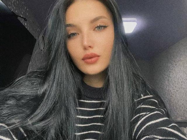AinkingMandarinka with long black hair, wearing a black and white striped sweater, has full red lips, eyeliner, and a neutral expression, in a dimly lit room.