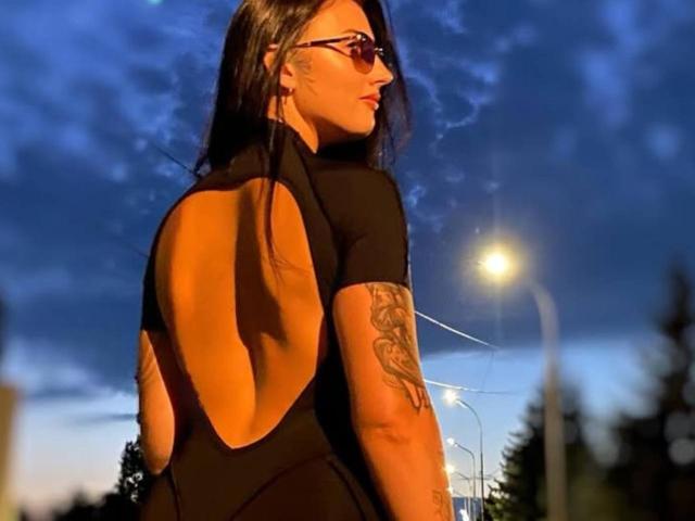 AlinkaMandarinka with long black hair, wearing sunglasses and a black backless top, showing a tattoo on her right arm, standing outside at dusk.