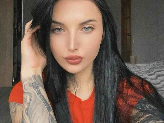 AlinkaMandarinka with long black hair, blue eyes, and red lipstick, wearing a red shirt, has a tattooed arm and is looking at the camera with a pouty expression.