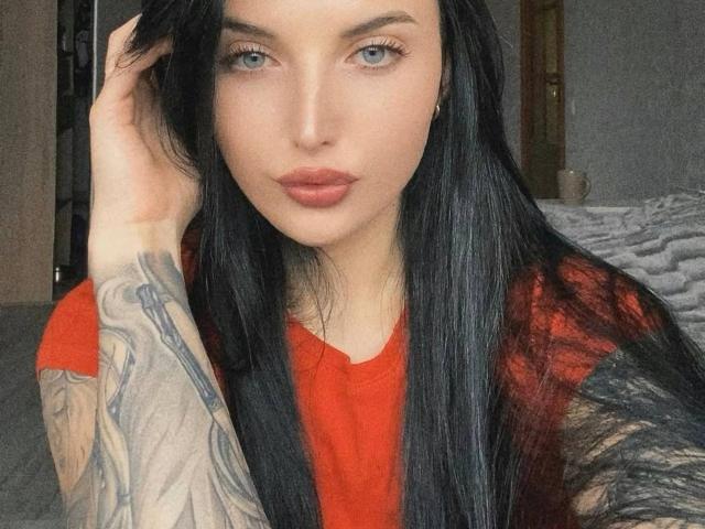 AlinkaMandarinka with long black hair, blue eyes, red shirt, and tattoos on her right arm, looking at the camera with her hand in her hair.