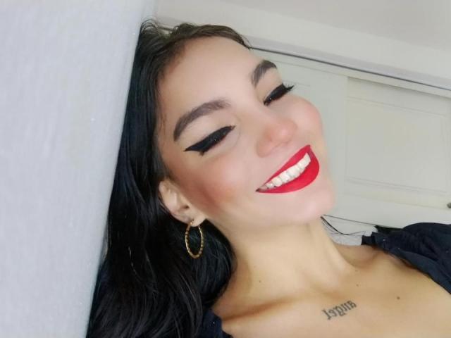 AurenLys con delineador negro, lápiz labial rojo, aretes de aro dorados, sonriendo, mostrando los dientes, cabello oscuro, tatuaje en el pecho, apoyada contra una pared blanca. Los senos ligeramente visibles..