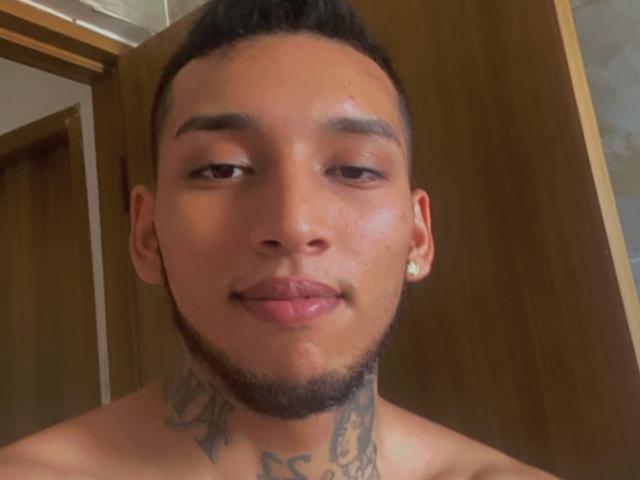 EvanVelvet with short black hair, light brown skin, tattoos on neck, small earrings, slight beard, sticking out tongue, shirtless, wooden door in background.
