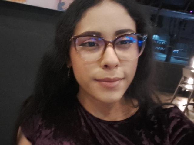 MarilynHills has dark hair, glasses, and a slight smile, wearing a purple velvet shirt, with her tits barely visible under the shirt. Her ass is not in the frame.