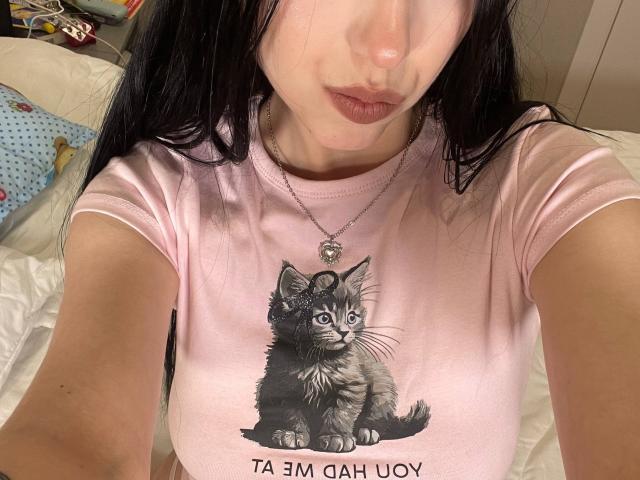 AmaiRin mit blasser Haut, dunklem Haar und einem pinken Hemd mit einem niedlichen grauen getigerten Kätzchen, trägt eine Halskette mit einem kleinen Anhänger und zeigt auf einem Selfie ihre leicht vorwitzigen Lippen..