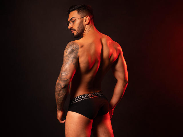 ThonyMuscle, muscular, tattooed, dark skin, short black hair, glasses, black underwear, red lighting, standing, back to camera, showing ass, looking over shoulder, dark background.