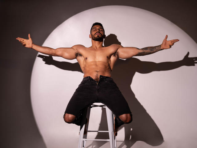 ThonyMuscle, shirtless, muscular, tattooed arm, black pants, sitting on white stool, arms out, pointing fingers, shadowed, light circle behind, dark background, short beard, confident pose.