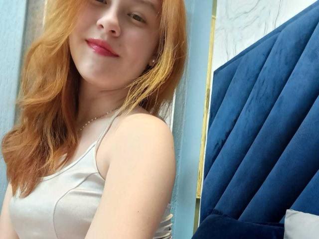 TaylorCarpert has red hair, fair skin, and wears a white tank top showing her small tits. She's smiling with pink lipstick, standing near a blue, velvet headboard.