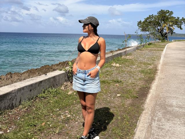 ThaliaAres with tan skin, black bra, and blue denim skirt, wearing a gray cap and black sneakers, standing by the ocean, hands in pockets, looking to the side.