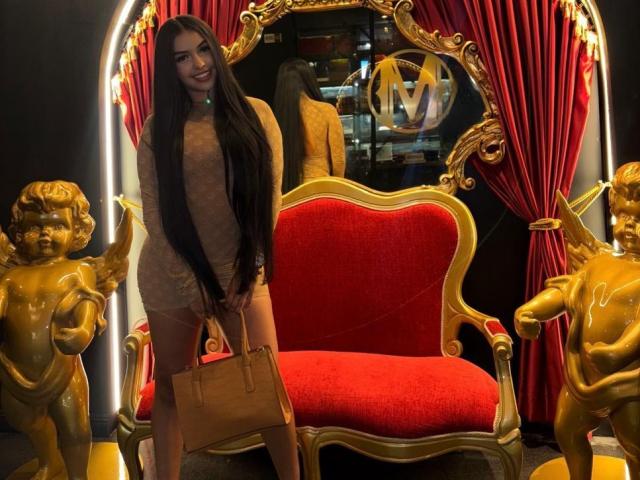 IvanaJovic stands with long black hair, wearing a sheer beige dress, holding a tan handbag, in front of a red velvet throne with gold cherub statues.
