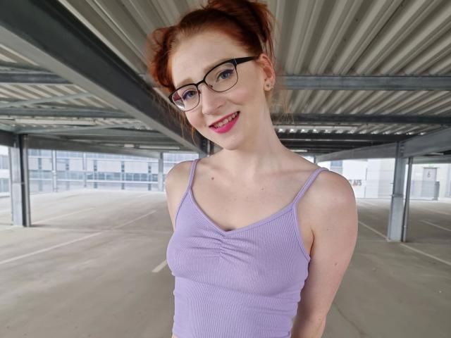 FinaFoxy-hot with red hair, glasses, and pink lipstick, wearing a purple tank top, showing her small tits, smiling under a metal roofed parking structure.