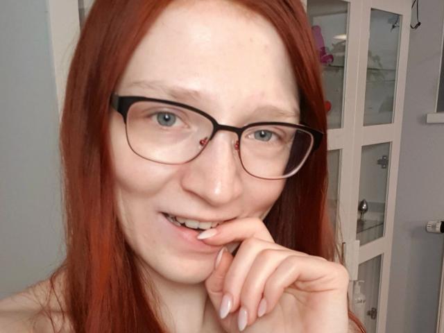 FinaFoxy-hot with red hair, glasses, pale skin, and a slight smile, biting her finger with a playful expression, showing her teeth and nails.
