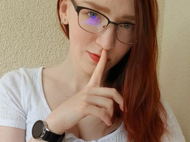 FinaFoxy-hot with red hair, glasses, white top, finger to lips, watch on wrist, purple digital face overlay, showing her tits and ass slightly through her top.