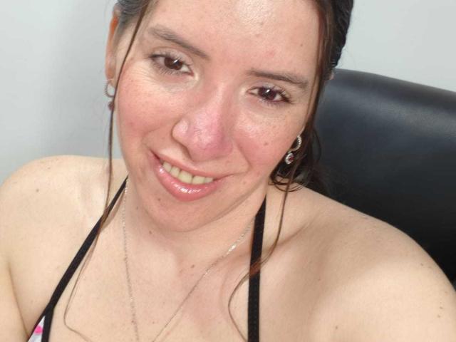 CoffeeCaress has a smiling face with light brown skin, small tits, brown eyes, and dark hair. She's wearing a black halter top, hoop earrings, and a thin necklace.
