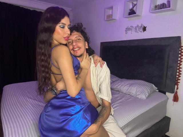 ArielleAndAxel, with long brown hair and blue dress, sits on bed, back to camera, showing ass. Axel, with curly hair, smiles, holding white towel, arms around Arielle.