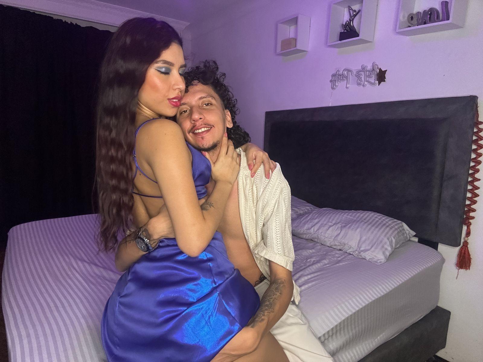 ArielleAndAxel, with long brown hair and blue dress, sits on bed, back to camera, showing ass. Axel, with curly hair, smiles, holding white towel, arms around Arielle.