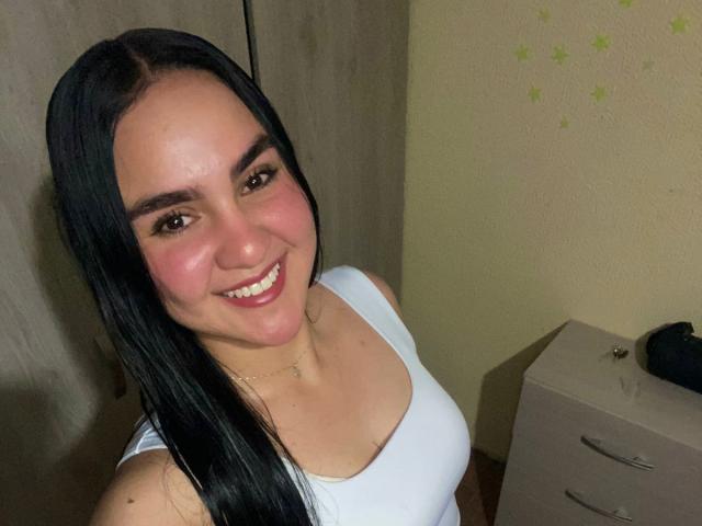 Valery Duran, com cabelos longos e pretos, sorrindo, vestindo uma regata branca, mostrando seus seios, com blush rosa nas bochechas, olhos castanhos e um colar de ouro em um selfie..