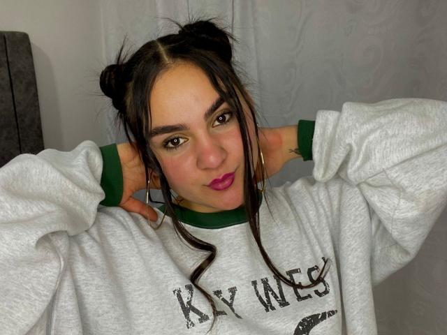 Valery Duran, portant un sweat gris gonflé, des poignets verts, deux chignons noirs, du rouge à lèvres rose, les mains derrière la tête, regardant la caméra, peau mate, yeux foncés, léger sourire, tatouage visible sur le bras gauche..