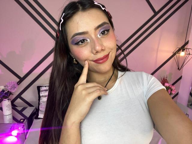 AlannaJonson with long brown hair, purple eyeshadow, red lipstick, white tee, finger on cheek, black necklace, pink headband, smiling, pink and purple lights in background.
