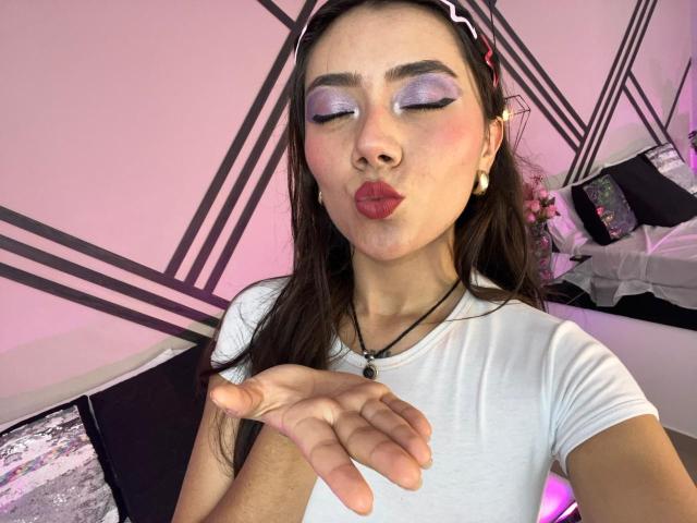 AlannaJonson with purple eyeshadow, red lipstick, and white tee, pouting her plump, glossy lips, hand near face, black headband, earrings, necklace, pink-purple lighting, and geometric background.