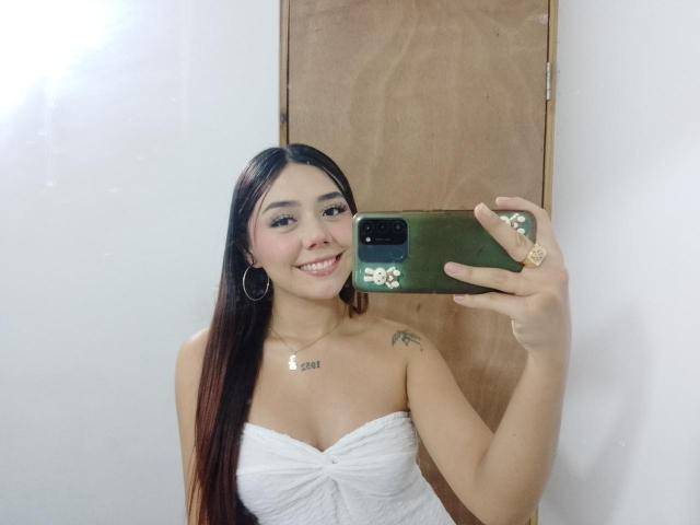 AlannaJonson with long brown hair, smiling, holding a green phone, wearing a white strapless top, showing small tits, tattoo on right shoulder, hoop earrings, and a necklace.