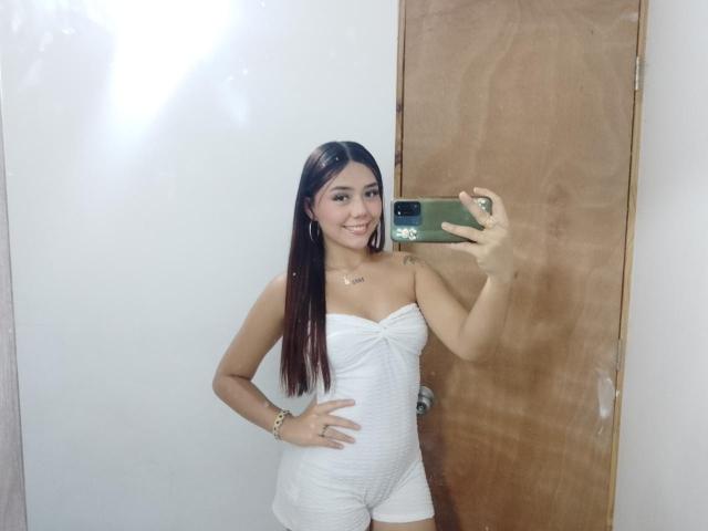 AlannaJonson with long brown hair, smiling, wearing a white strapless dress, holding a green phone, standing in front of a wooden door, showing her small tits and slim ass.