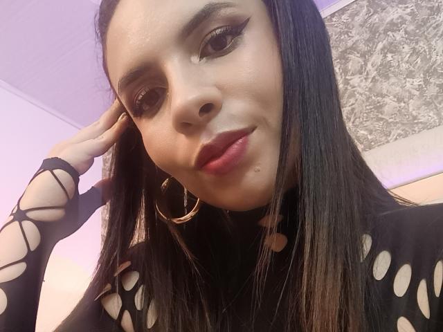 VioletaStone with long black hair, wearing a black fishnet top, gold hoop earrings, and red lipstick, looking at the camera with a slight smile. Her hand touches her head.
