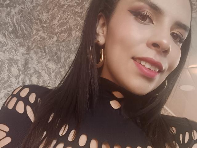VioletaStone with long black hair, wearing a black, cut-out top, gold hoop earrings, and pink lipstick, smiling at the camera. Her brown eyes are accentuated with winged eyeliner.