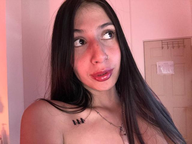 Sophie Beaumont tem cabelos pretos longos, um piercing no nariz e lábios vermelhos brilhantes. Seus seios pequenos são visíveis, com uma tatuagem preta no ombro direito. Iluminação rosa suave..