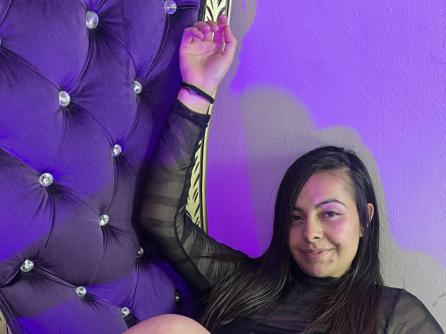 BonnieRosses with black hair, smiling, wearing a sheer black top, arm raised, touching a purple, tufted, diamond-studded headboard, under purple lighting.