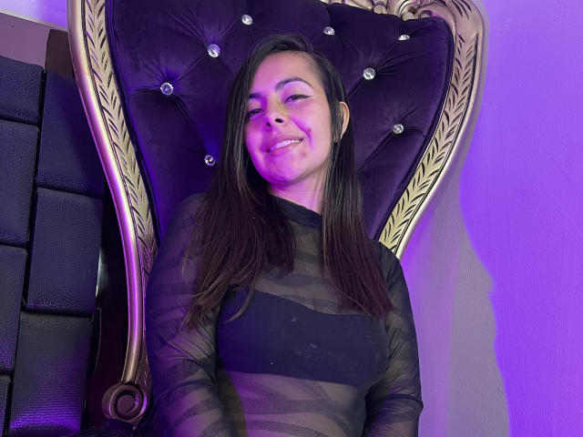 BonnieRosses with dark straight hair, smiling, sitting on a tufted, purple velvet chair, wearing a sheer black long-sleeve top, purple light in the background.