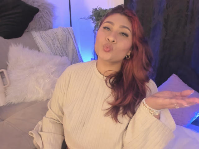 ElyRoss with red hair, pouty lips, and gold hoop earrings, wearing a beige ribbed sweater, hand up, looking cute. Boobs not visible. Soft blue light in background.