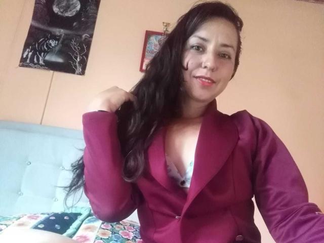 DouglasAndVioleth with long black hair, wearing a maroon blazer, showing a bit of blue lace bra, smiling, touching her hair, pink lipstick, and light brown skin.